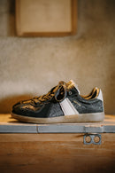 Black and gold sneaker with a white sole on a wooden surface, Obroi