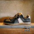 Black and gold sneaker with a white sole on a wooden surface, Obroi