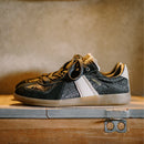 Black and gold sneaker with a white sole on a wooden surface, Obroi