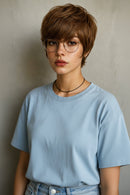 A close-up portrait of a person with a short, layered brown wig, wearing round metal-framed glasses, a black cord necklace, and a light blue crewneck shirt against a neutral gray background, Obroi