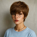 A close-up portrait of a person with a short, layered brown wig, wearing round metal-framed glasses, a black cord necklace, and a light blue crewneck shirt against a neutral gray background, Obroi