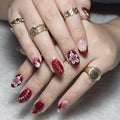 Close-up of hands with red nail polish and floral designs, wearing gold rings on a neutral background, Obroi