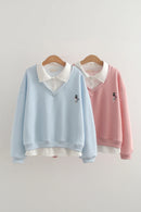 Two long-sleeve V-neck sweatshirts hanging on wooden hangers against a plain light background. The front sweatshirt is light blue with a white collar and a small black cat embroidery on the chest, Obroi