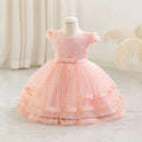 A toddler girl's formal party dress displayed on a mannequin. The dress features a light pink honeycomb patterned bodice with short puff sleeves and a matching pink tulle tiered skirt with gold lace trim, Obroi