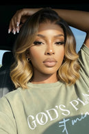 A woman wearing a shoulder-length wavy wig with dark brown roots and blonde highlights, posing in a car while wearing an olive green graphic t-shirt, Obroi