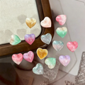 A set of small heart-shaped decorative pieces in pastel gradient and glitter finishes, arranged on a light surface with a wooden frame edge visible, Obroi
