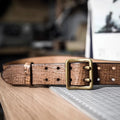 A brown crocodile-embossed leather belt with a square antique brass buckle, resting on a wooden surface next to a cutting mat, Obroi