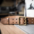 A brown crocodile-embossed leather belt with a square antique brass buckle, resting on a wooden surface next to a cutting mat, Obroi