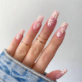 Hand with pink floral nail design wearing gold rings on a light gray background, Obroi