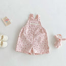 A baby girl's pink floral overall romper laid flat on a light gray surface, styled with white baby shoes, a small card, and a pink plush bunny keychain, Obroi