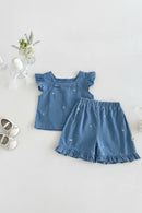 A baby girl's two-piece outfit set consisting of a blue denim-like top with ruffled sleeves and matching shorts with ruffled hems, both decorated with tiny colorful embroidered flowers. The outfit is displayed in a styled flat-lay on a light gray textured background, surrounded by decorative props including white baby shoes, a candle, a small flower vase, and a pearl hair clip, Obroi