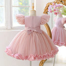 A light pink toddler girl's party dress displayed on a dress form mannequin, featuring puff sleeves, a matching pink bow at the waist, and a ruffled tulle skirt, with a mirror in the background reflecting the dress, Obroi