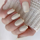 A close-up of a hand with glossy almond-shaped nails painted in a warm off-white/cream shade, each finished with a thin gold accent at the tip, against a white knit sleeve, Obroi
