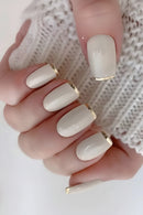 A close-up of a hand with glossy almond-shaped nails painted in a warm off-white/cream shade, each finished with a thin gold accent at the tip, against a white knit sleeve, Obroi