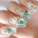 Close-up of a hand with coffin-shaped nails featuring a clear and mint green ombre design, decorated with white dried flowers and tiny gold glitter details, Obroi