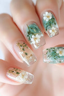 Close-up of a hand with coffin-shaped nails featuring a clear and mint green ombre design, decorated with white dried flowers and tiny gold glitter details, Obroi