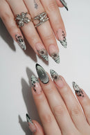 A close-up of a hand with long, embellished almond-shaped nails. The nails feature a mix of pale pink, glossy black and white zebra-like patterns, cat-eye gradient tips, and silver rhinestone floral accents, Obroi