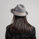Person wearing a gray fedora hat with a feather on a plain background, Obroi