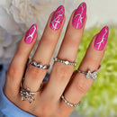 Hand with pink nail art and multiple silver rings against a blurred floral background, Obroi