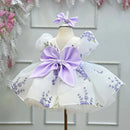 White dress with purple floral patterns and a large bow on a mannequin against a decorative background.