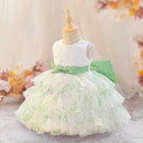 A sleeveless toddler girl's party dress displayed on a wooden mannequin. The dress features a white lace bodice, a light green satin waist sash with a bow, and a tiered ruffled skirt in white with a light green floral toile pattern, Obroi