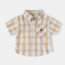 A baby boy's short-sleeve button-up shirt in a beige, white, and yellow plaid pattern, featuring a chest pocket with a small cartoon character patch, Obroi