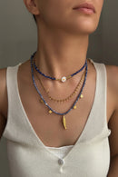 Necklace with blue and gold beads worn by a person in a beige top, Obroi