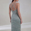 A woman shown from the back wearing a sleeveless, floor-length evening gown covered in shimmering sequins and fringe. The dress has thin crisscross strappy details across the open back, Obroi