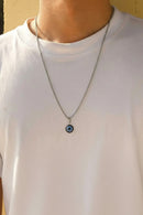 Person wearing a necklace with an eye-shaped pendant on a plain background, Obroi