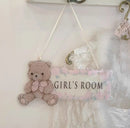 A hanging decorative sign for a girl's room featuring a teddy bear on the left and the words 'GIRL'S ROOM' on the right, with a pink floral border and white ribbon cords for hanging, Obroi