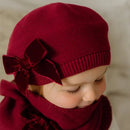 Child wearing a red knit hat with a large bow on a neutral background, Obroi