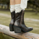 A pair of dark brown leather ankle boots with block heels and side gore details, worn with white socks while sitting on a wooden log in an outdoor park setting, Obroi