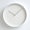 A minimalist round wall clock with a matte white face and frame, black hands, and a small gold center accent, photographed against a clean white background, Obroi