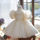 A cream-colored toddler girl's party dress displayed on a white mannequin dress form. The dress features a square neckline, voluminous puff sleeves, a beaded bodice, and a tiered tulle skirt, Obroi