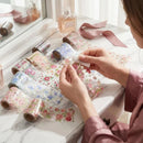 Person working with floral tape on a desk with various items including perfume bottles and ribbons, Obroi