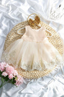 A baby girl's sleeveless tulle dress in pale blush pink and cream, decorated with gold lace embroidery and a pearl-embellished neckline, displayed on a woven straw mat with a gold bow headband, pink flowers, and a tiara, Obroi