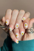 Close-up of manicured fingernails with a daisy design: white petals, yellow centers, and small smiley faces, shown on a person's hands with silver rings, Obroi