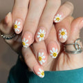 Close-up of manicured fingernails with a daisy design: white petals, yellow centers, and small smiley faces, shown on a person's hands with silver rings, Obroi