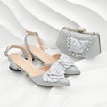 A pair of silver glitter pointed-toe slingback flats with layered fabric flowers on the toes, shown alongside a matching silver glitter clutch bag with a rhinestone-embellished chain closure, set against a white wavy background, Obroi