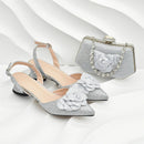 A pair of silver glitter pointed-toe slingback flats with layered fabric flowers on the toes, shown alongside a matching silver glitter clutch bag with a rhinestone-embellished chain closure, set against a white wavy background, Obroi
