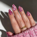 A close-up of a hand with almond-shaped nails painted in a pink and white zebra stripe pattern, wearing a gold ring and a pink knit cuff, set against a dark gray background with a soft shadow, Obroi