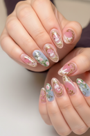 A close-up of a hand with manicured nails featuring a pastel pink and light blue floral design with gold foil accents, Obroi