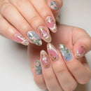 Close-up of manicured fingernails with a glossy pink and pale blue floral design, gold foil accents, and small pink flamingo motifs, shown on hands against a soft light background, Obroi