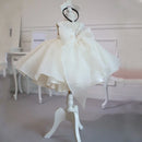 A white baby girl's dress displayed on a white mannequin stand in an indoor room. The dress features a sweetheart neckline with delicate lace trim, a large central bow, and a voluminous layered tulle skirt, Obroi