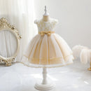 A toddler girl's formal party dress displayed on a white dress form mannequin. The dress features a cream-colored lace bodice with gold embroidery, short puff sleeves, a matching cream tulle skirt, a beige satin waistband with a large bow, and gold lace trim along the hem, Obroi
