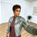 A woman taking a mirror selfie wearing a sage green satin bomber jacket with brown stripes over a ribbed burgundy dress, in a bright room with light wood floors and a potted plant, Obroi