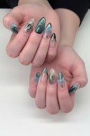 A close-up of a hand with long almond-shaped nails decorated in a fantasy-inspired teal, green, and silver nail art design. The nails feature iridescent teal sections, silver filigree, and clear sections with white line art, Obroi