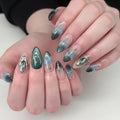 A close-up of a hand with long almond-shaped nails decorated in a fantasy-inspired teal, green, and silver nail art design. The nails feature iridescent teal sections, silver filigree motifs, and clear sections with silver star details, Obroi