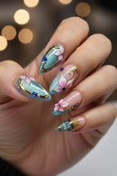 A close-up of a hand showcasing elaborate almond-shaped nail art. The design features a gradient of light blue and pink with metallic gold foil details, 3D pink flowers, blue jewel accents, and white pearl embellishments, Obroi