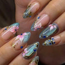 A close-up of a hand showcasing elaborate almond-shaped nail art. The design features a gradient of light blue and pink with metallic gold foil details, 3D pink flowers, blue jewel accents, and white pearl embellishments, Obroi